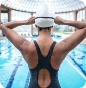 A swimmer seen from the back adjusting their swim cap while standing at the edge of an indoor swimming pool.
