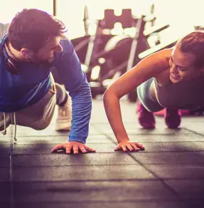 A man and a woman doing push-ups together in a gym, both smiling and engaging in a friendly competition.