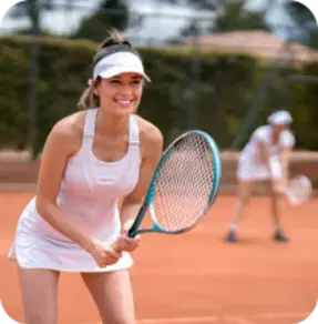 A smiling woman in a white tennis outfit holding a racket on a clay tennis court with another player in the background.